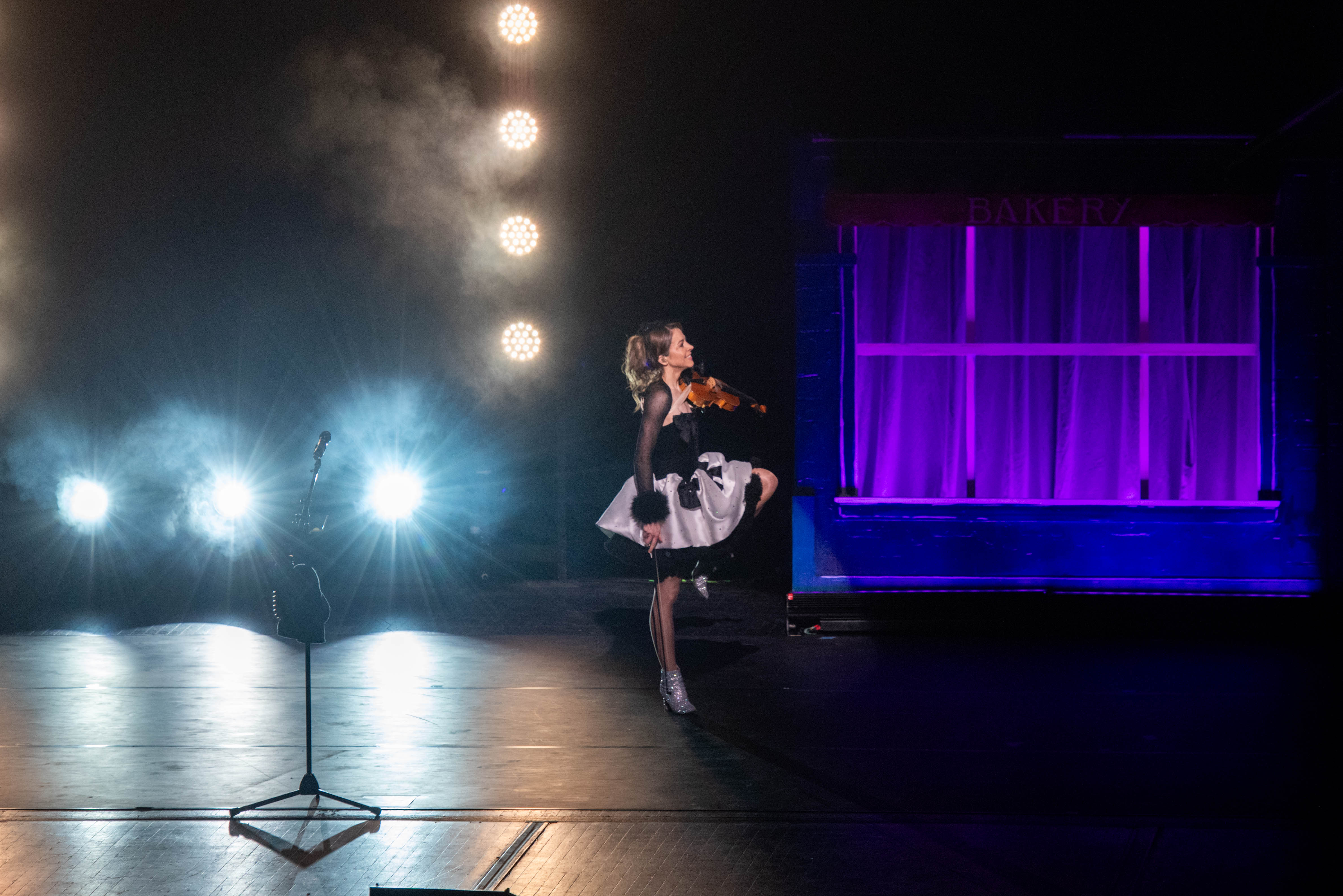 Lindsey Stirling in a spotlight plays violin on stage, dressed in a black and white outfit, balanced on one leg, with dramatic lighting and purple backdrop