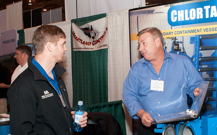 Two men converse at a trade show booth. The man on the left holds a water bottle, wearing a black jacket. The booth displays equipment and banners.
