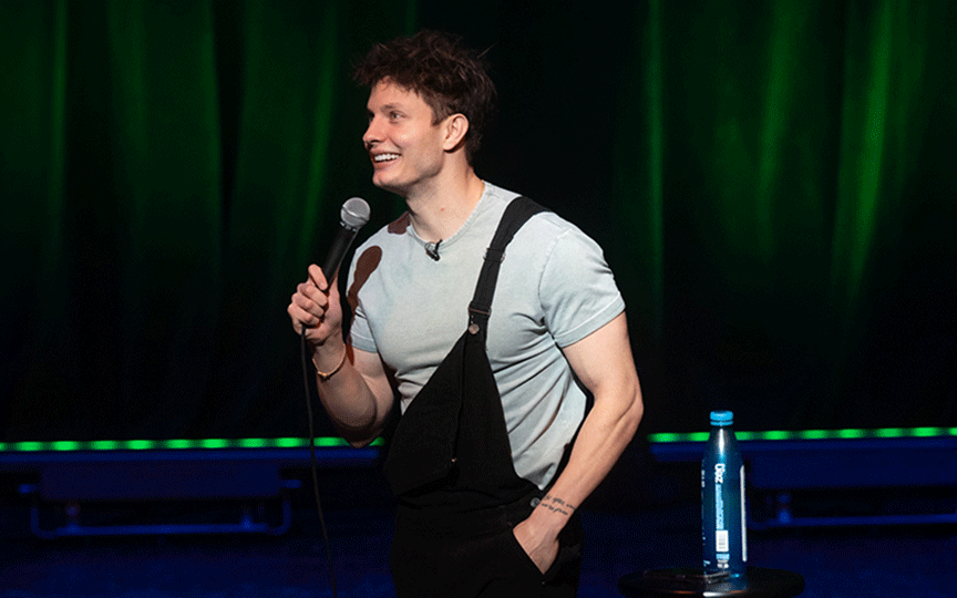 Matt Rife on stage wearing a light shirt and black overalls, holding a microphone and smiling. A spotlight highlights him against a dark green curtain with lime green floor lights.