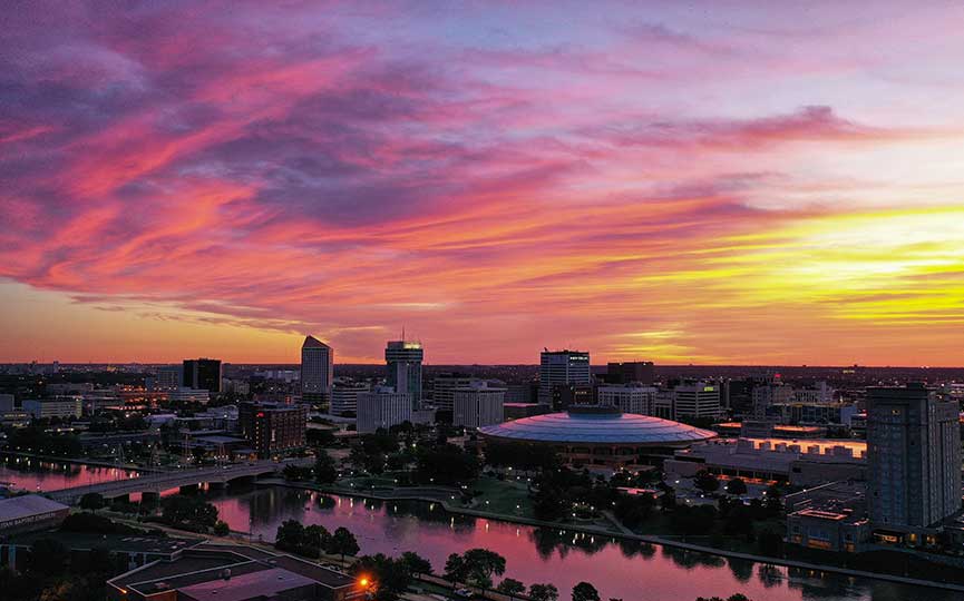 A city skyline at dusk with a vibrant sunset in the background. The sky displays hues of pink, orange, and purple, reflecting on a river below. Century II is featured in the center.