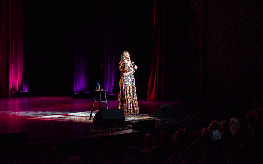 Leanne Morgan on stage in a patterned dress, under purple and red lights, speaks into a microphone. A stool with a water bottle beside her; an audience watches.
